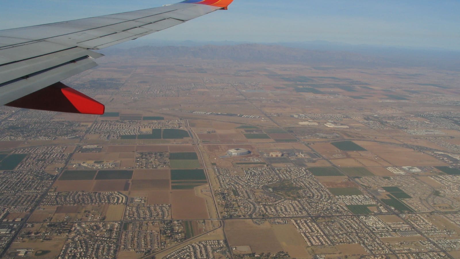 An aerial view from an airplane window over Glendale, Arizona, showing the city's grid of suburbs and farmland with the domed stadium visible in the distance