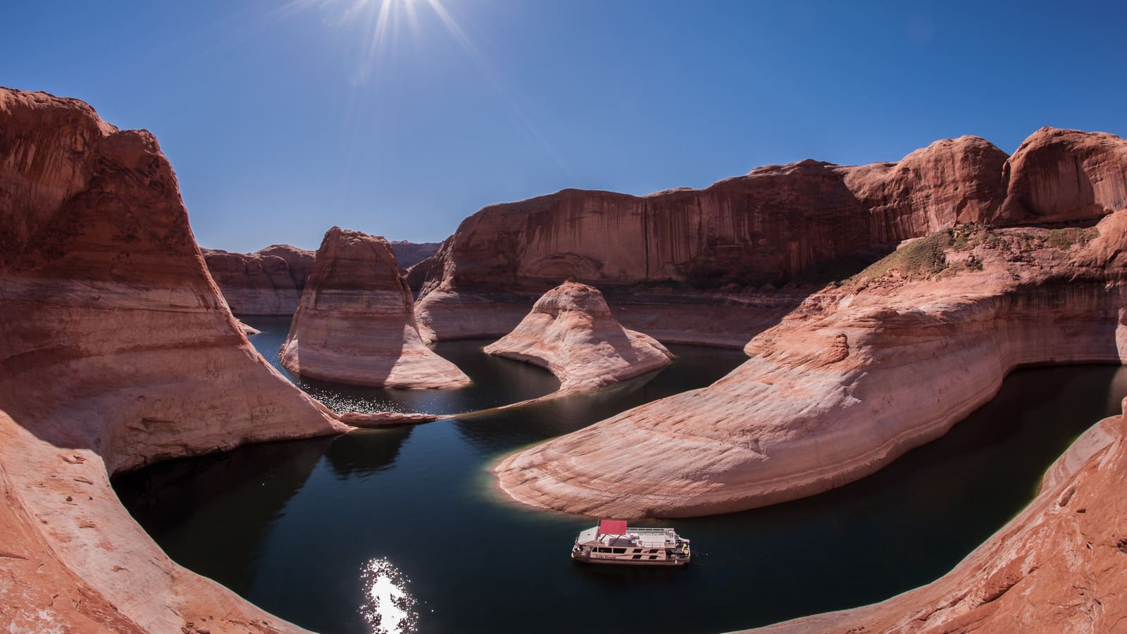A houseboat floats on Lake Powell's green water beneath the dramatic curving red-rock walls of Reflection Canyon